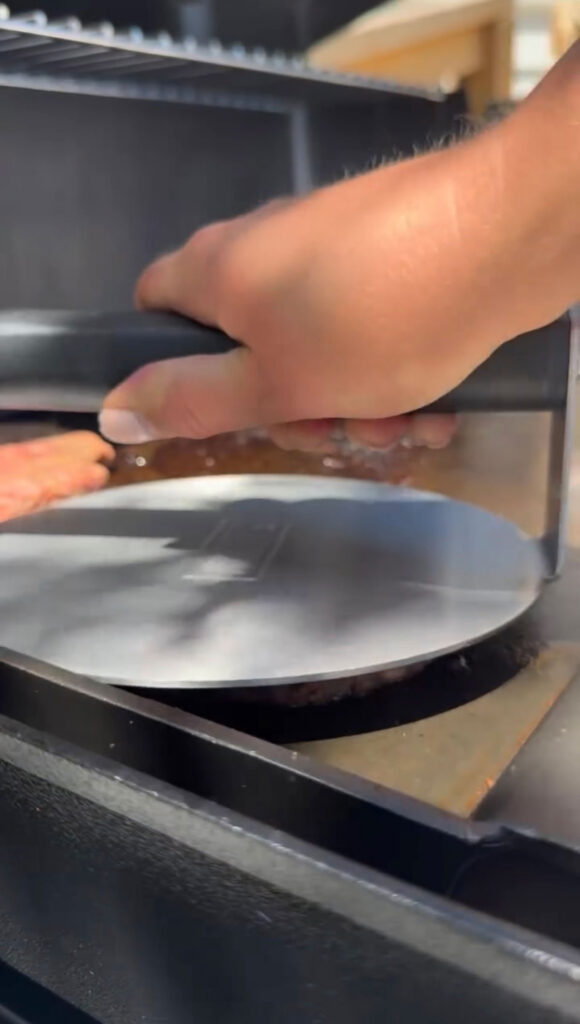 A hand smashing the pickle smash burger down with a press on a griddle.