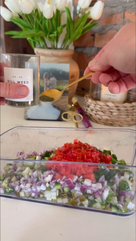 A hand adding dill to pickle salsa in a plastic container.