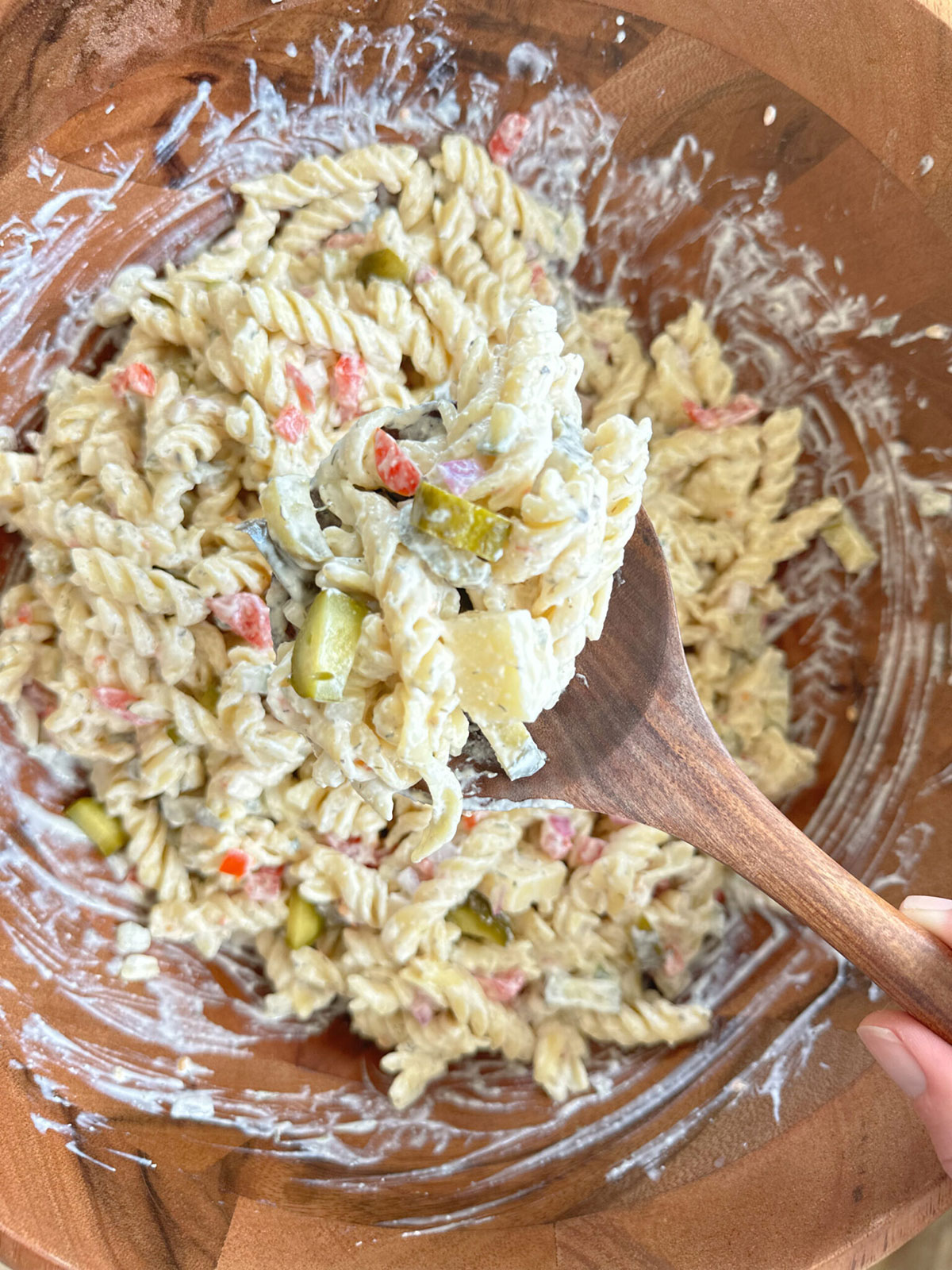 A glass bowl with a wooden spoon picking up a spoonful of dill pickle pasta salad.