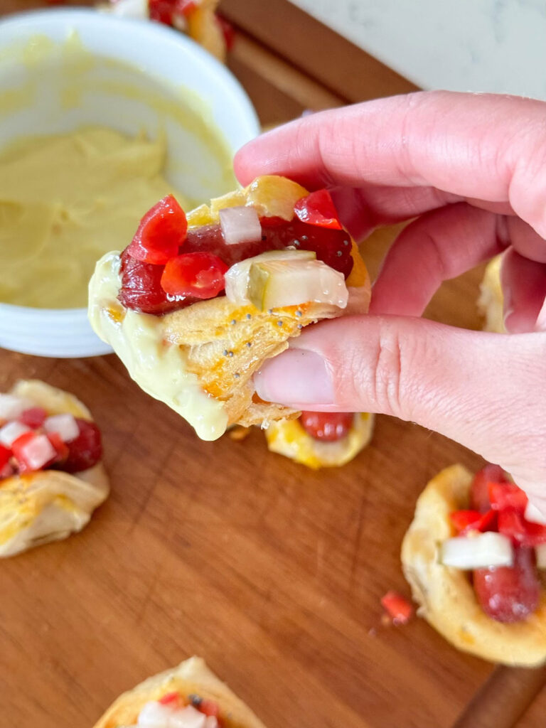 A hand holding a mini hot dog with more on a cutting board below.