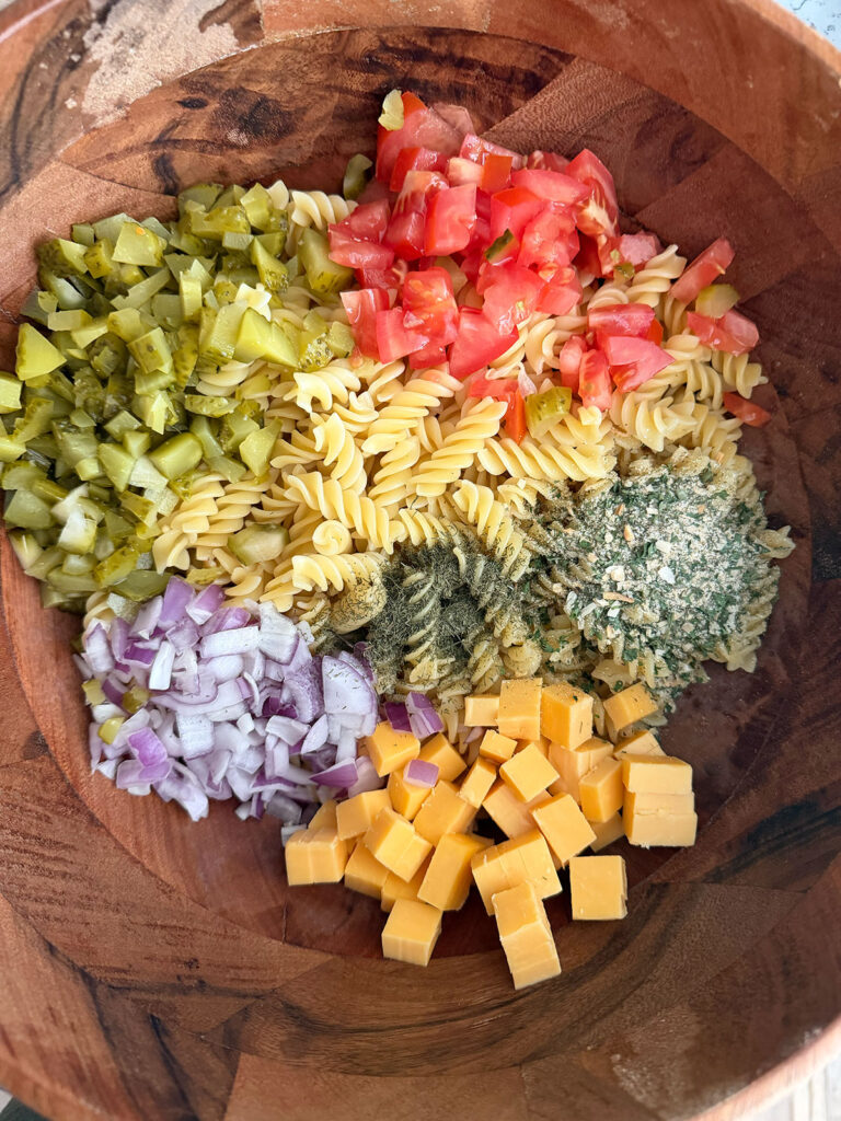 A wooden bowl with piles of ingredients for dill pickle pasta salad.