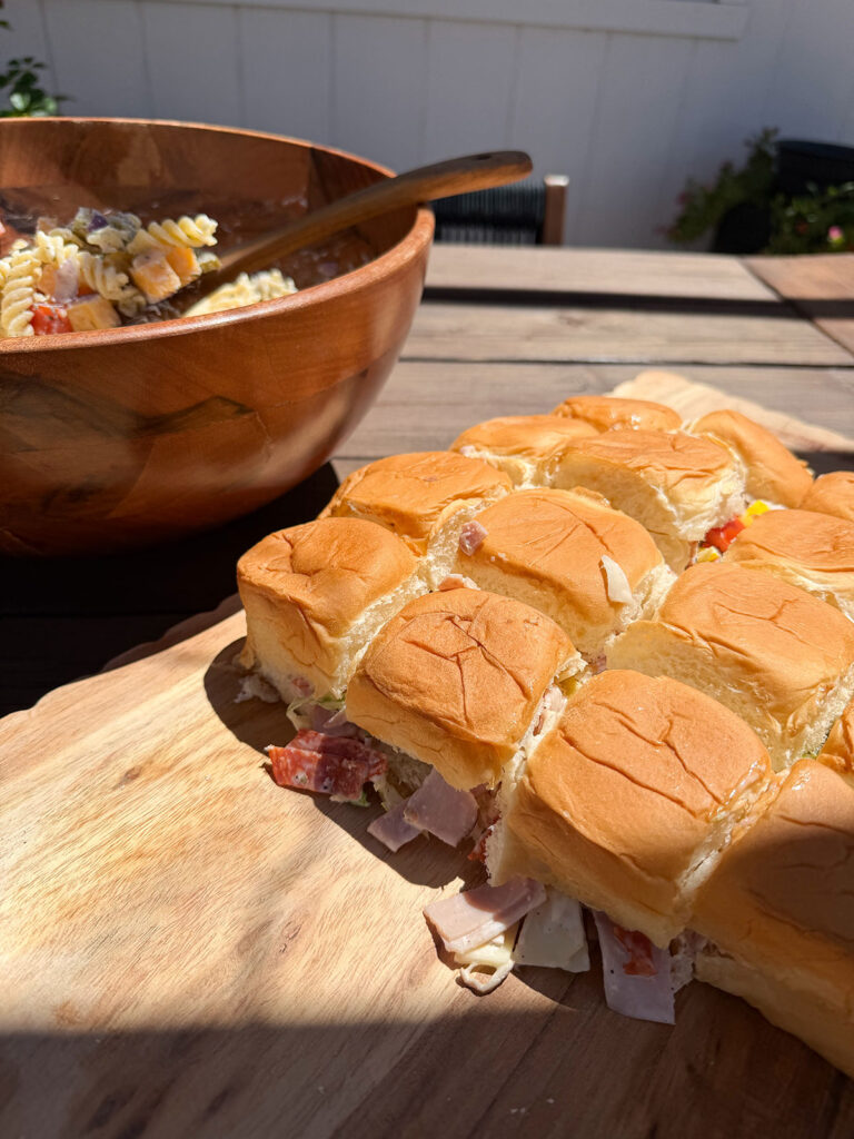 Chopped Italian sliders on a wooden board with a wooden bowl of pasta salad in the background.