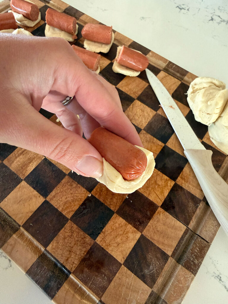 mini hot dogs-4 A hand wrapping a mini hot dog with a piece of biscuit dough. A checkered cutting board with more ingredients is below.