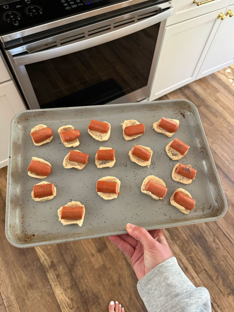 A hand holding a baking sheet with mini hot dogs ready to go into the oven.