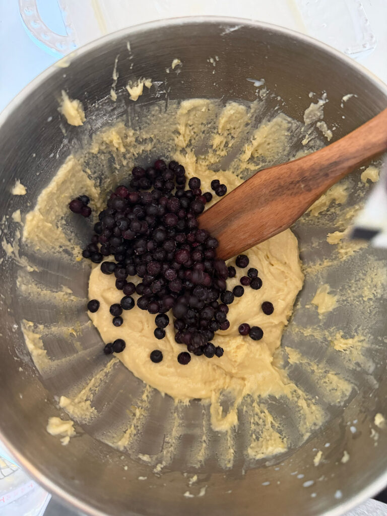 A stand mixer bowl with cake batter and frozen blueberries on top. A wooden spoon is resting on the side.