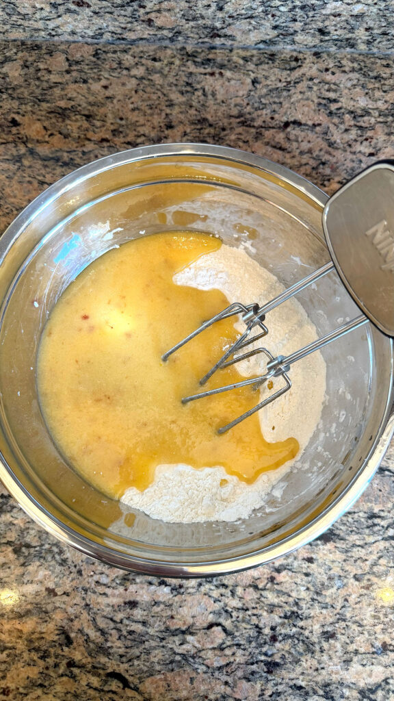 A metal bowl with liquid and dry ingredients being mixed with a hand mixer for strawberry bundt cake.