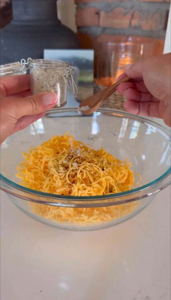 hand holding container of seasoning and adding to clear bowl with shredded cheddar cheese and ingredients in it