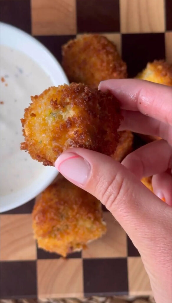 hand holding whole fried pickle cheese ball with fried pickle cheese balls underneath and dip in white bowl.