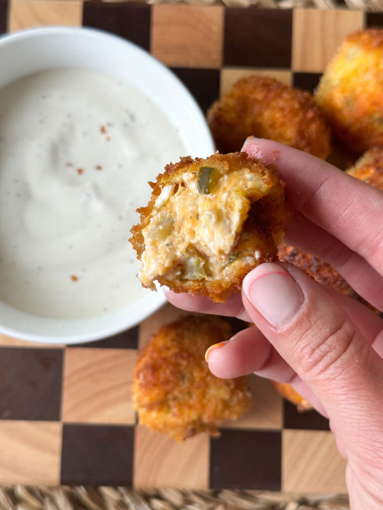 Hand holding opened up fried pickle cheese ball with bowl of dip in white bowl underneath. pile of fried pickle cheeseballs on cutting board under, too.