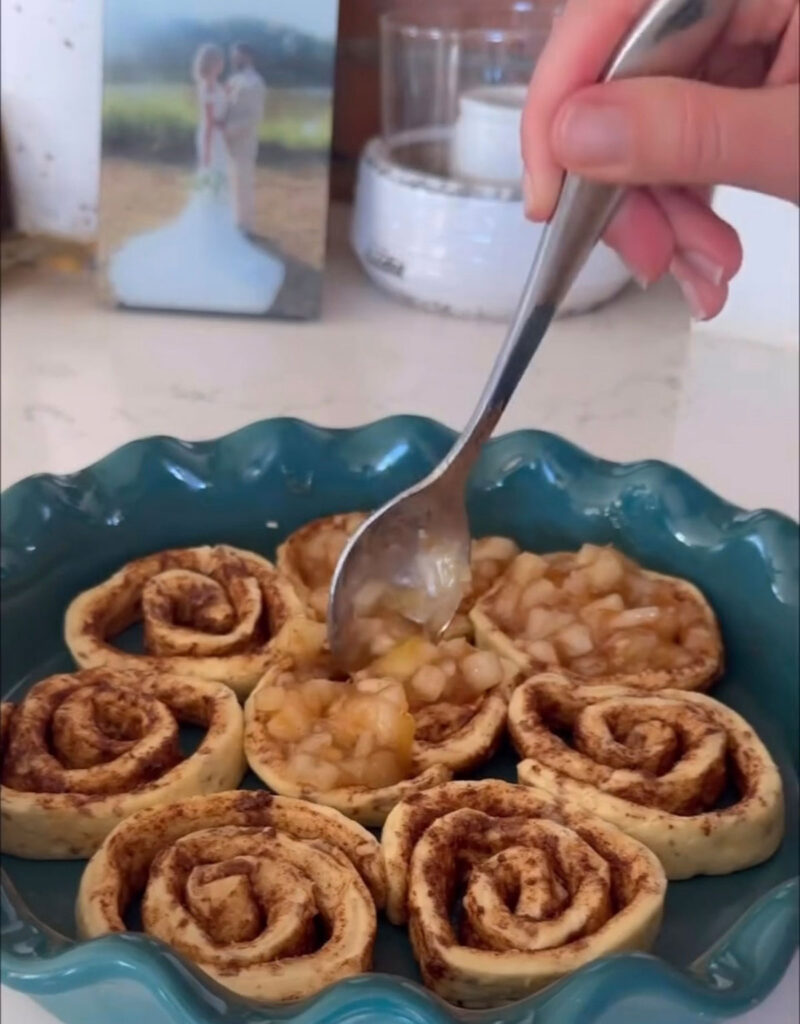 apple pie cinnamon rolls in pie plate with spoon adding apple pie filling on top