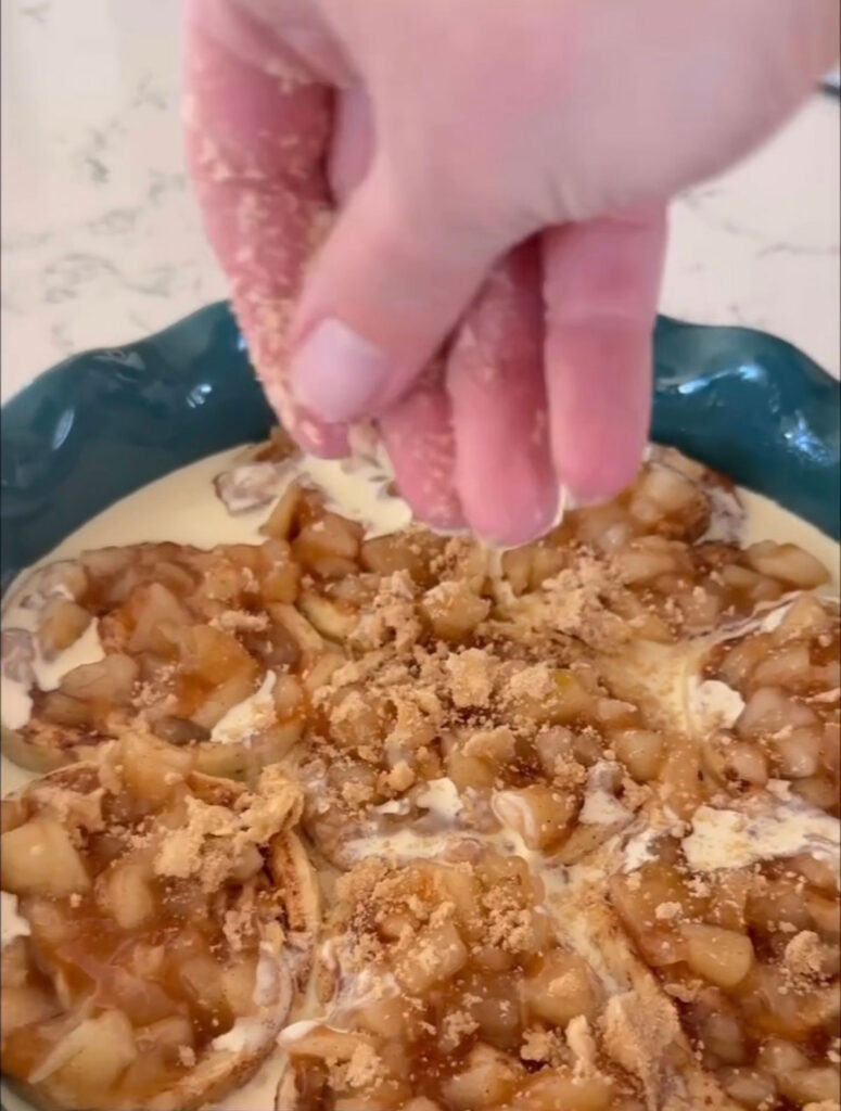pie plate with apple pie cinnamon rolls uncooked in it, with hand sprinkling on topping