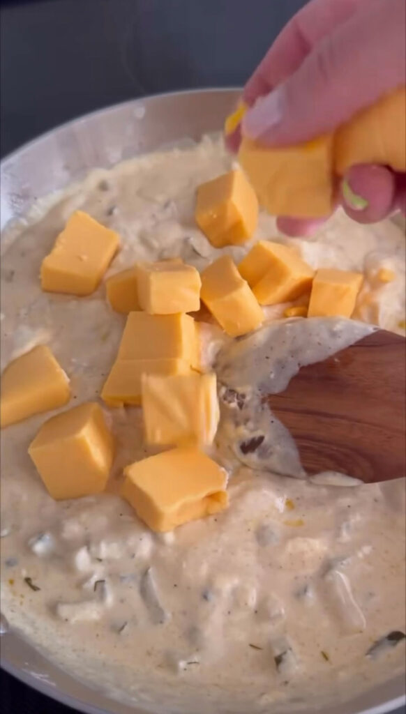 hand dropping chunks of cheese into pan with creamy mixture in it