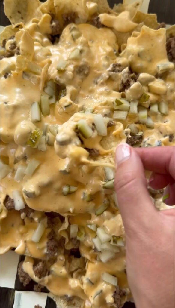 hand holding a chip above pile of nachos covered with toppings including nacho sauce