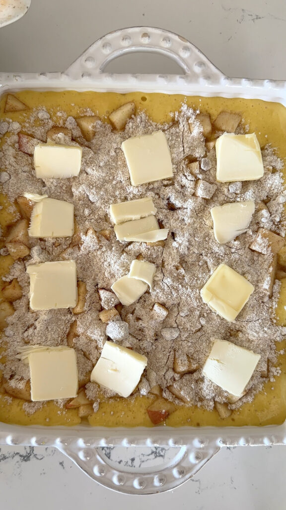 apple pie cake batter with diced apples, crumb topping and pats of butter on top ready to be baked
