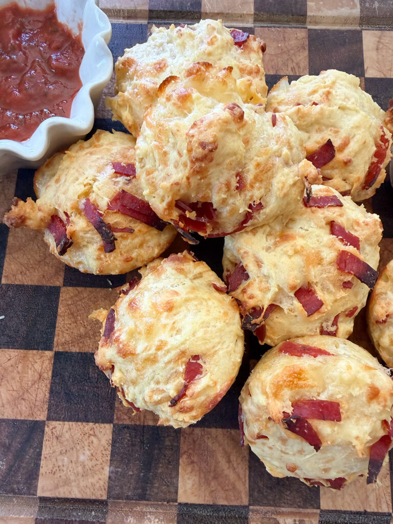 pile of baked pizza muffins on cutting board next to white bowl of marinara sauce