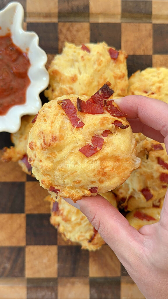 Hand holding pizza muffin over baked pizza muffins next to bowl of marinara sauce