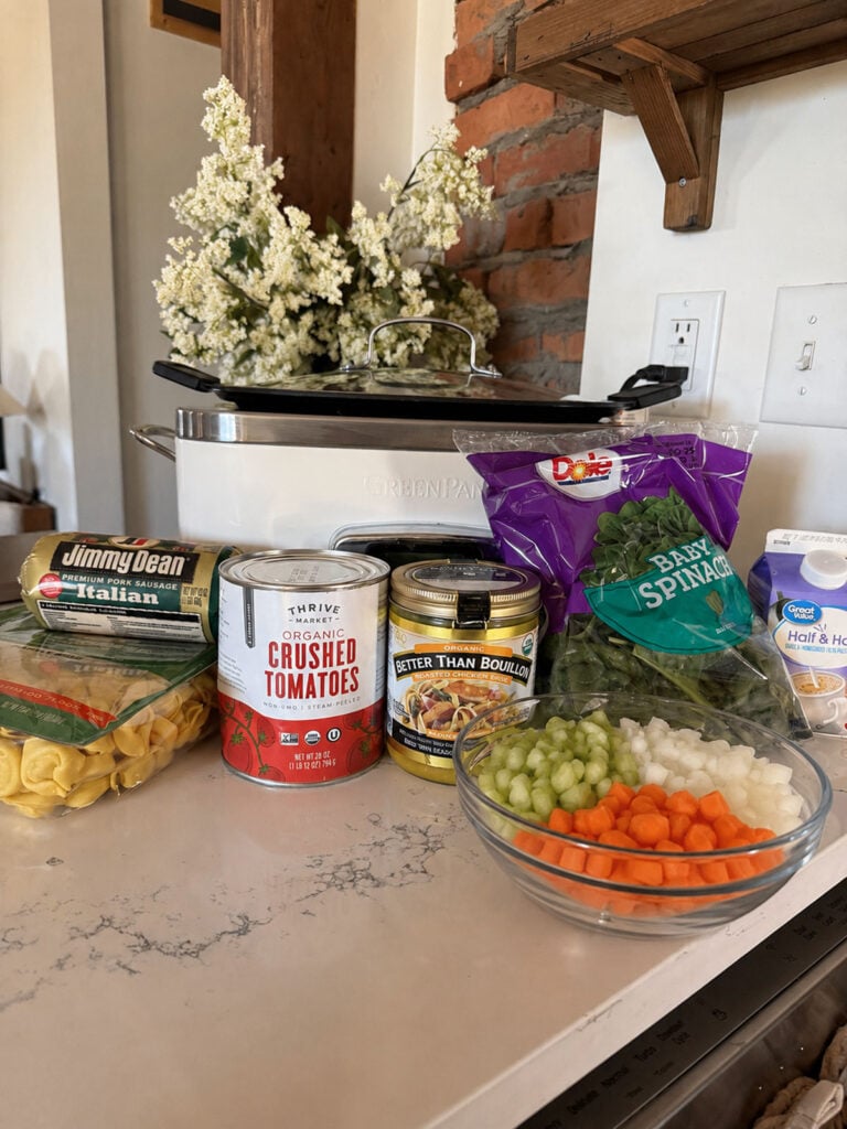 creamy sausage tortellini soup ingredients on counter with slow cooker in background and vase of flowers