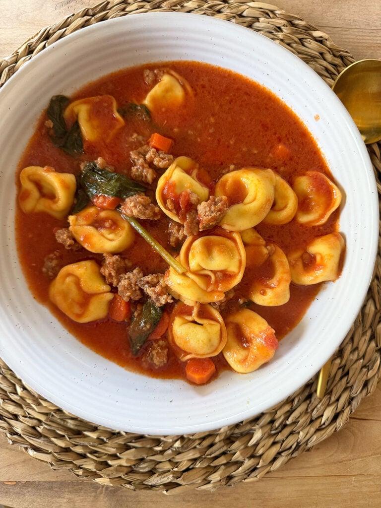 white bowl of sausage tortellini soup on placemat with gold spoon next to it