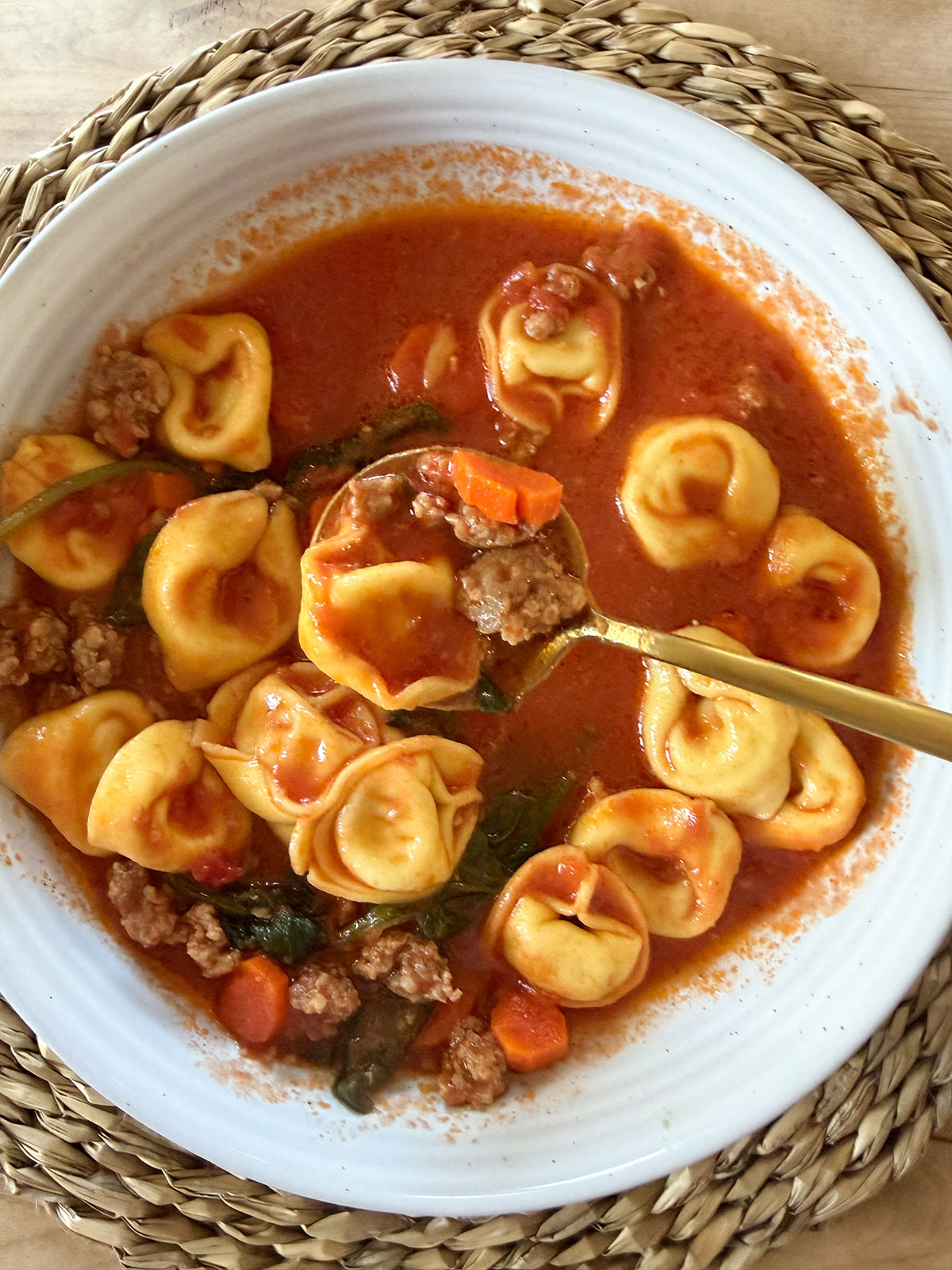 sausage tortellini soup in white bowl on placemat with gold spoon in it