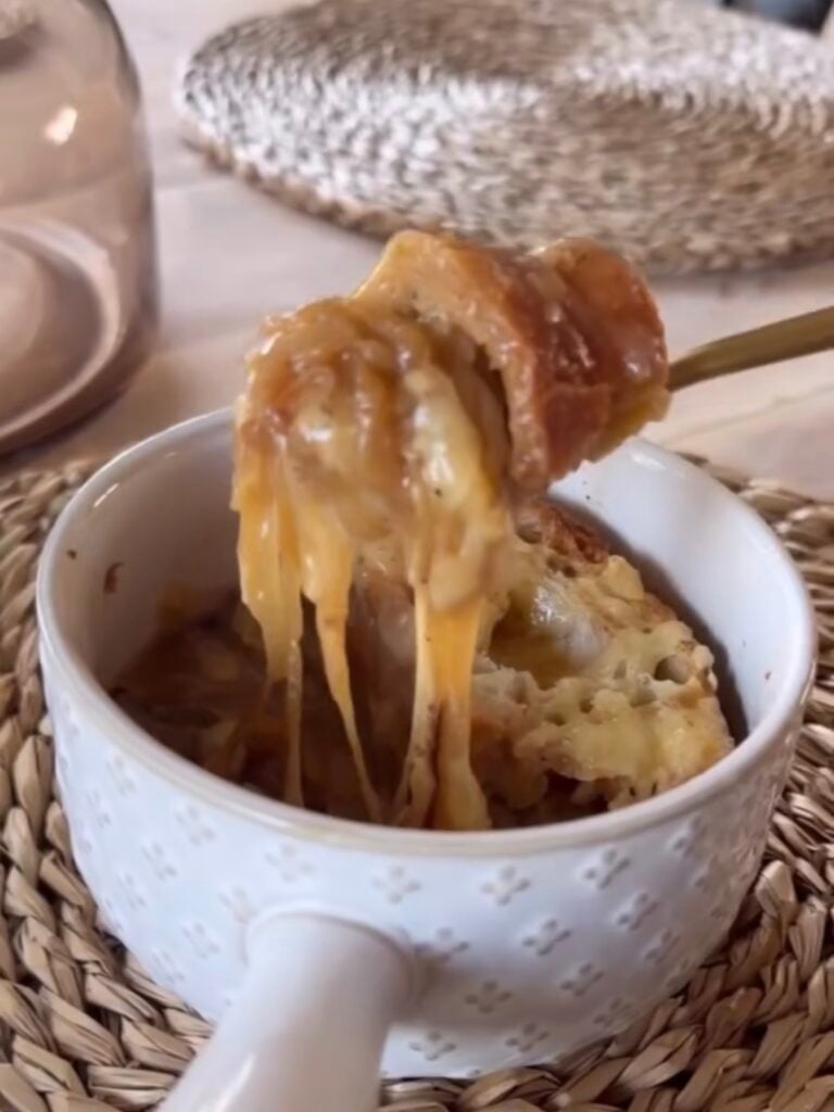 french onion soup in white crock with gold spoon. placemat in background